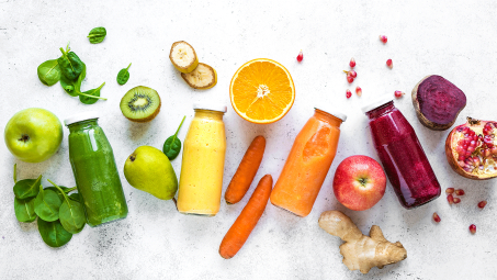 glass drink bottles filled with green, yellow, orange, and dark red liquids surrounded by fresh whole fruits and vegetables including spinach, kiwi, bananas, carrots, apples, pears, ginger, beats and pomegranate 