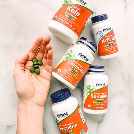 light skinned hand holding 6 green tablets next to an array of NOW supplements