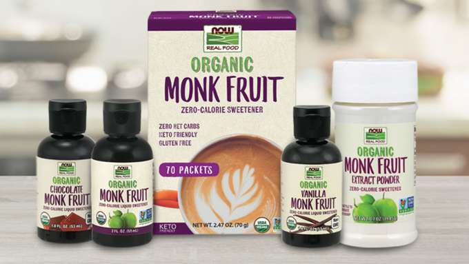 five Monk Fruit products on light colored counter - three small bottles of flavored liquid, one box of packets, one bottle of powdered Monk Fruit - all with light tan label with purple and green accents and lettering