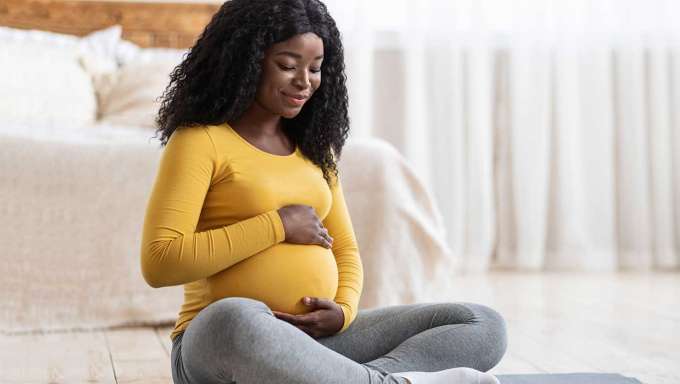 black woman in yellow shirt sitting cross legged on bedroom floor holding her pregnant stomach