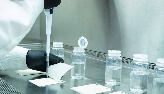 two white coated arms with black gloved hands working to test clear liquid on white paper with glass jars