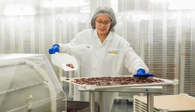 medium skin female presenting person wearing lab coat and blue gloves shoveling red softgels onto a metal tray