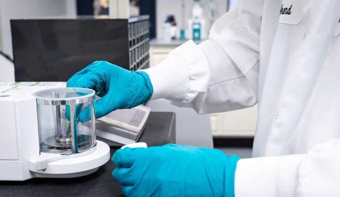 two white coated arms with blue gloved hands working to test clear product inside a glass jar