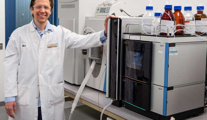 light skin male presenting person standing in a white lab coat next to a machine in a lab setting