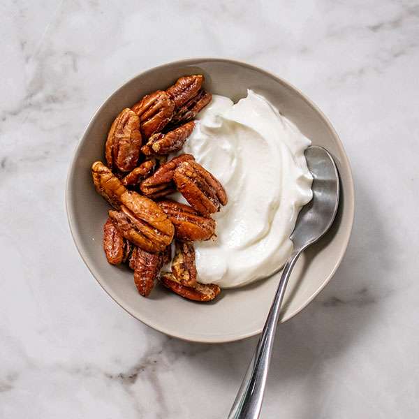 overhead view of candied pecans in a bowl with yogurt on one side sitting on a white marble counter