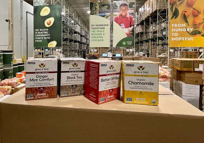 four new NOW tea boxes sitting on top of a closed box in a well lit warehouse with racks in the background