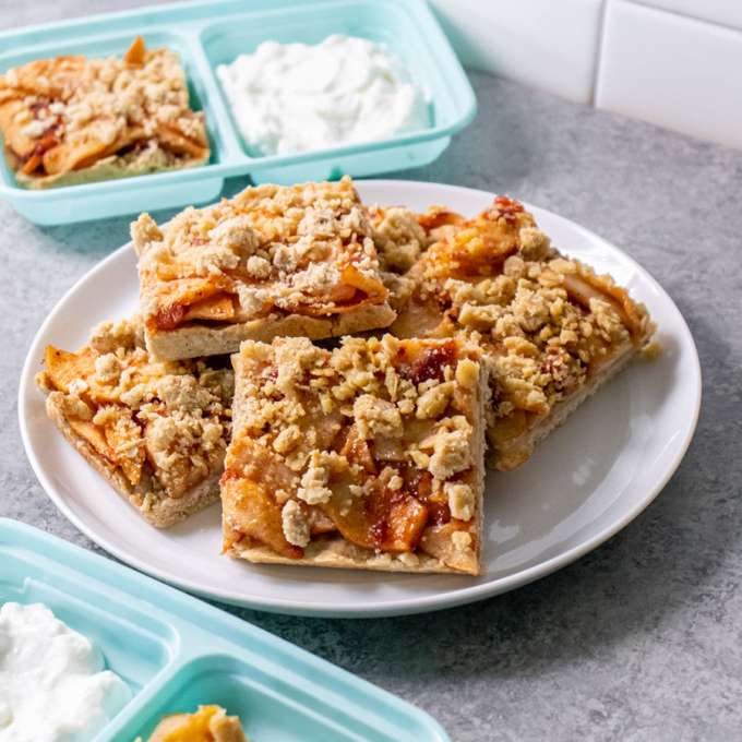 white plate with five square apple pie bars and two trays with bars and yogurt on them