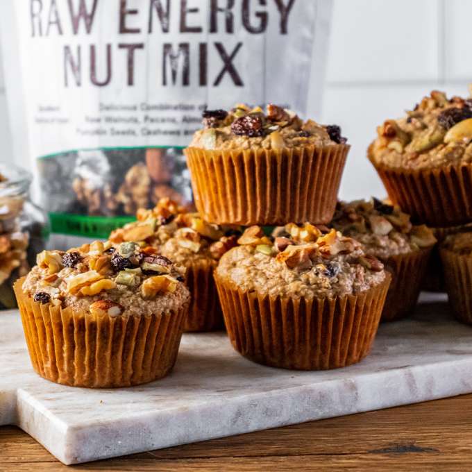 pile of Oat Bran Good Morning Muffins with a blurred bag of NOW Raw Energy Mix and jar containing its contents as well