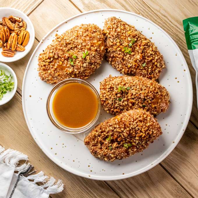 top view of Pecan Herb Crusted Chicken with maple dijon sauce on a white plate next to a bag of NOW Organic Pecans