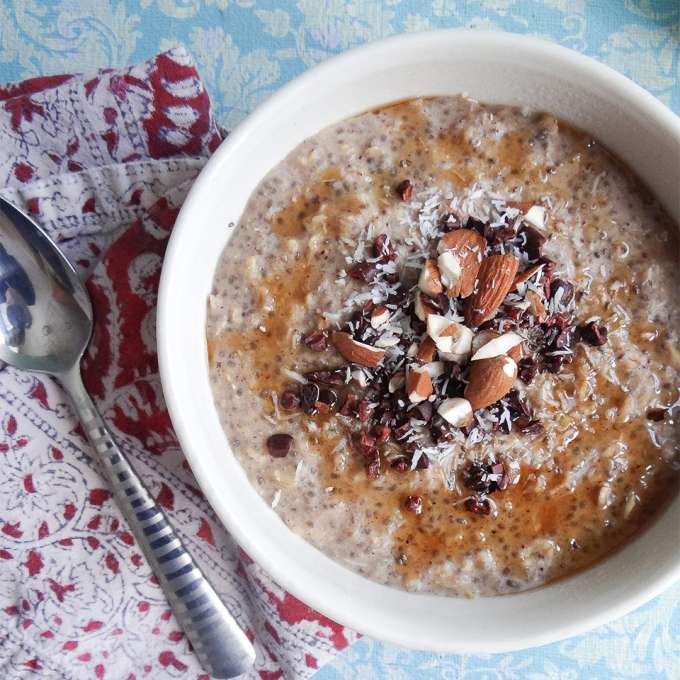 White bowl of oatmeal with a spoon resting on the left red and white batik placemat on a light baby blue and white floral leaf pattern tablecloth. The ingredients in the recipe are nicely displayed in the bowl showing oatmeal with specks of chia seed with a circular drizzle of maple syrup and toppings in the center of chopped almonds, cacao nibs and shredded coconut.