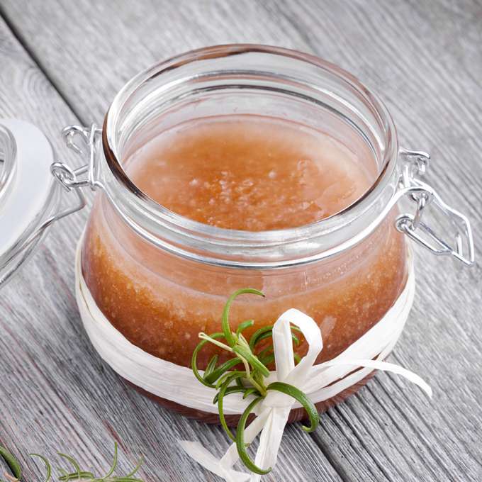 Short glass jar about three inches with white gasket and metal hinged top with a white ribbon wrapped around several times then tied in a simple bow with a green plant sprig tucked in the bow. Jar contains combined recipe ingredients that are amber in color and looks slightly coarse and the lid is open. The jar sits on a gray plank tabletop.