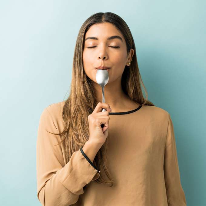 woman standing with eyes closed with tip of a large spoon in her mouth like she's eating a popsicle