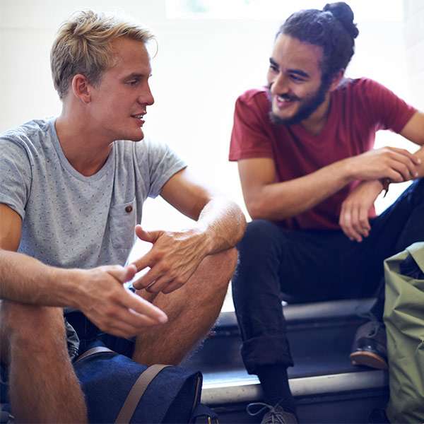 Three male-presenting people sit in a stairwell, relaxed and smiling, chatting to each-other.