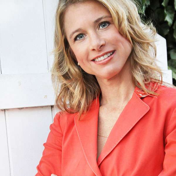 Wendy Bazilian smiles and looks at the camera outdoors while wearing a red blouse.