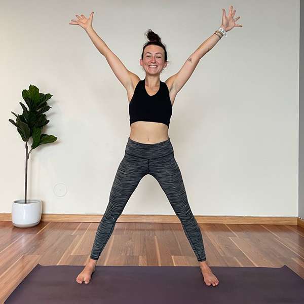 presenting as female wearing dark gray yoga pants and a black sports top exercising on a yoga mat in a white room with wood floors