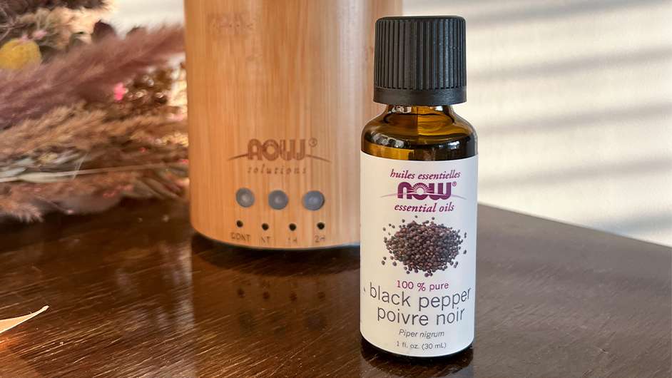 NOW Black Pepper Essential Oil bottle in front of a NOW Bamboo diffuser on a brown table with dried flowers in the background
