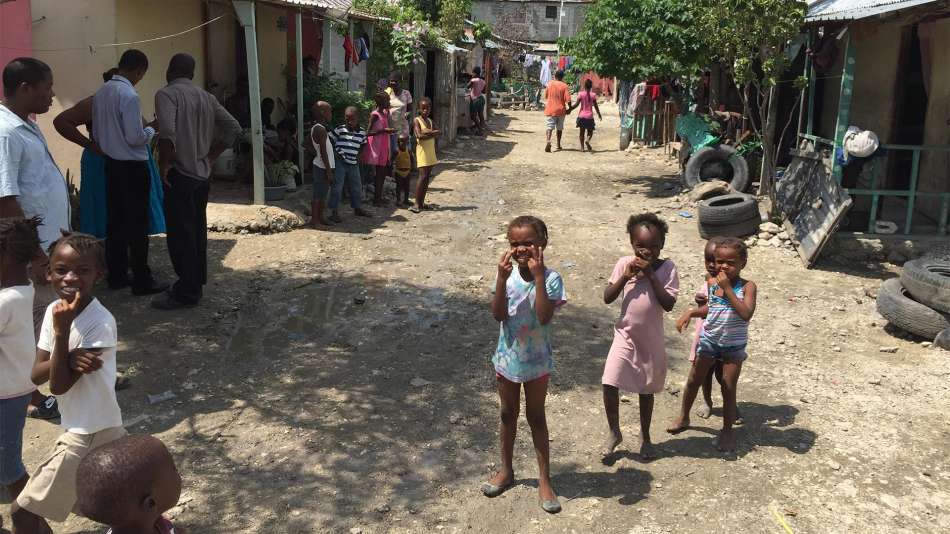 Three small dark-skinned children stand on a dirt road with buildings and other dark-skinned children and people lining the street.