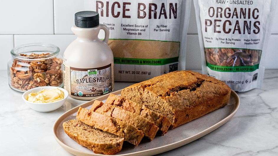 a loaf of rice bran banana nut bread in front of NOW syrup, rice bran, and pecans