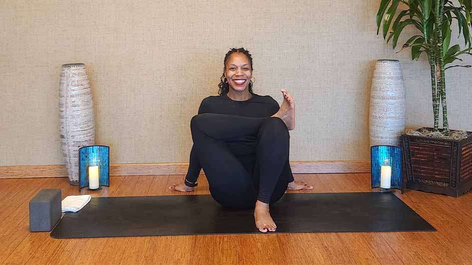 dark skin female presenting person sitting on a black yoga mat stretching her hips